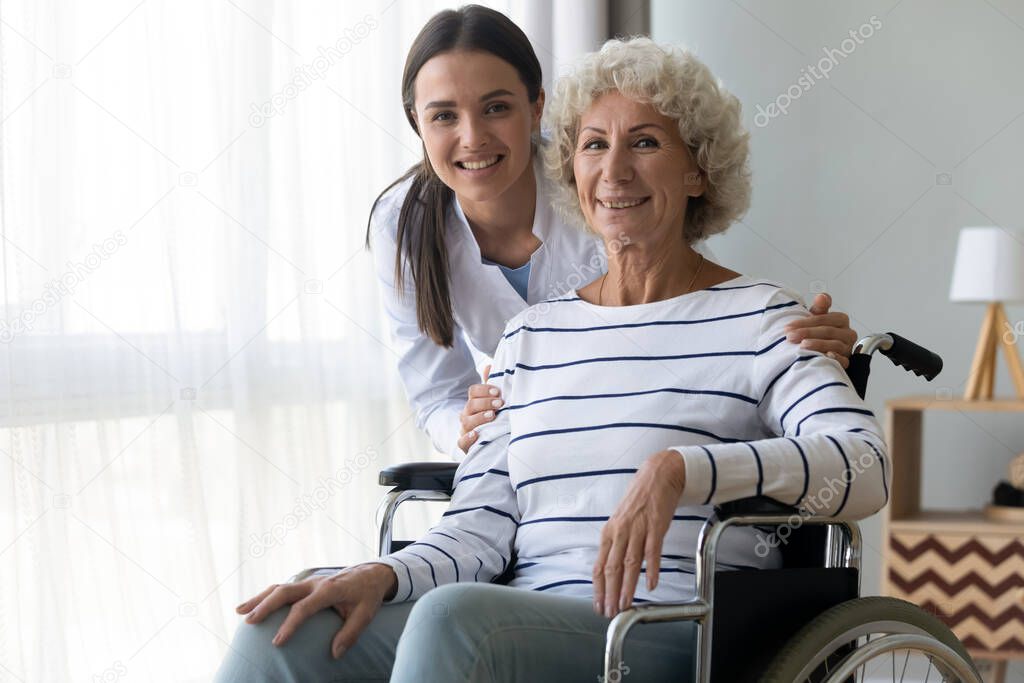 Caring nurse hug elderly disabled woman in wheelchair posing indoors