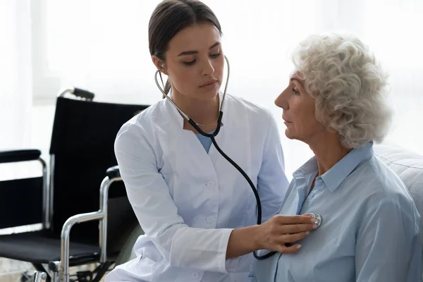 Nurse uses stethoscope listens hearts sounds of elderly disabled woman
