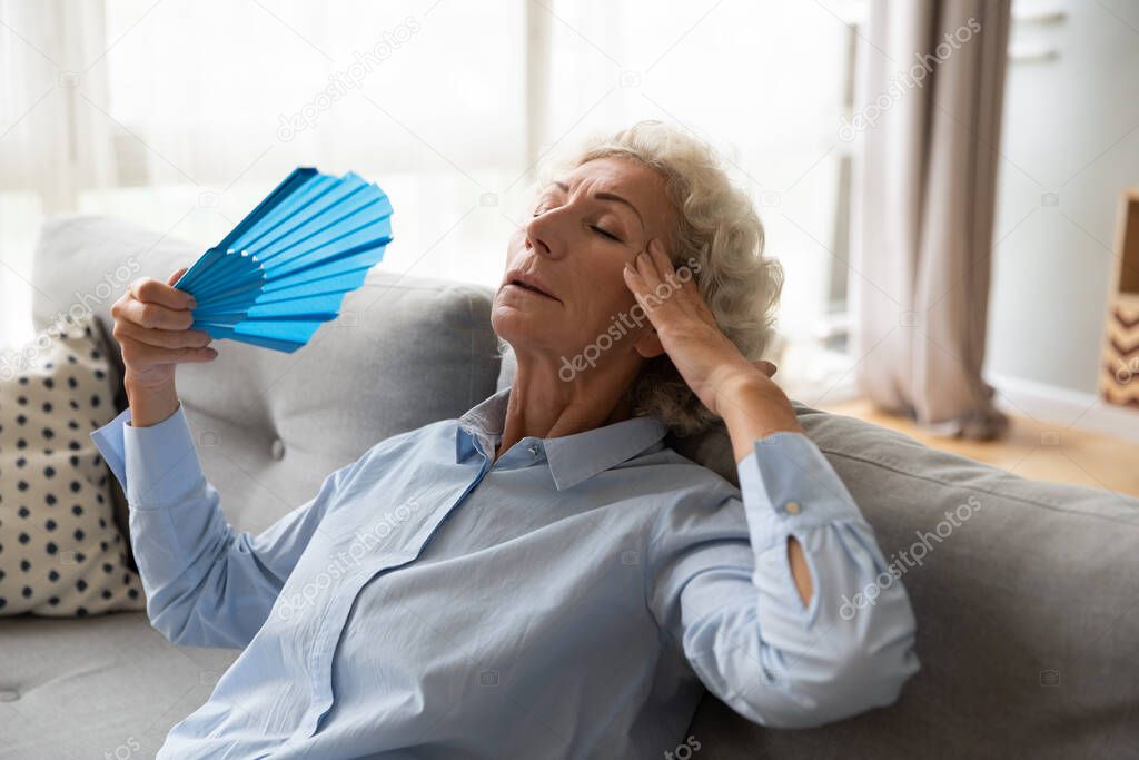 Overheated elderly female leaned on couch refreshing herself with fan