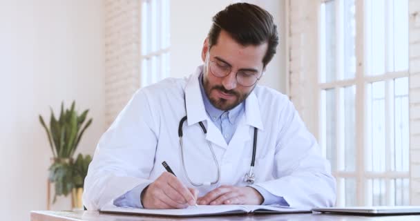 Gericht jonge mannelijke huisarts doet papierwerk op het kantoor van de kliniek. — Stockvideo