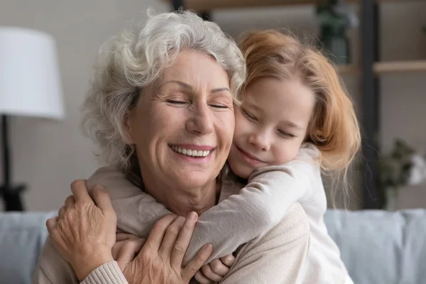 Заботливая маленькая девочка обнимает счастливую зрелую бабушку дома — стоковое фото