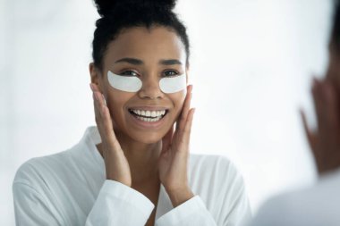 Ayna yansıması gülümseyen Afrikalı Amerikalı kadın hidrojel göz bantları uyguluyor.