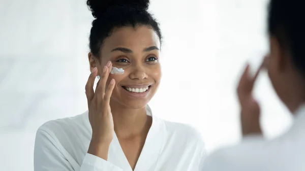 Riflessione specchio sorridente donna afroamericana applicando crema idratante — Foto Stock