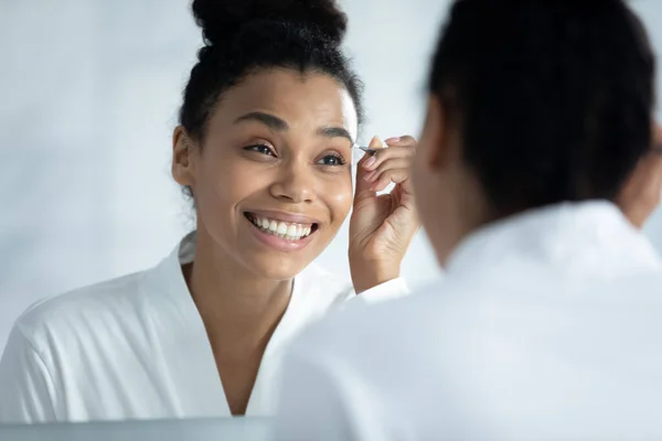 Aynanın yansıması gülümseyen Afrikalı Amerikalı kadın cımbızla kaş yoluyor. — Stok fotoğraf