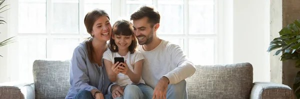 Parents and daughter sit on couch have fun using smartphone