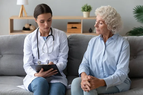 Young female caregiver listen writing health complaints of elderly woman — Stock Photo, Image