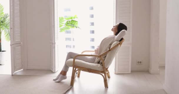 Glückliche junge Frau tagträumt in kuscheligem Stuhl neben riesigem Fenster. — Stockvideo
