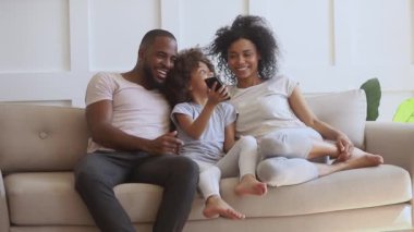 Happy african parents with kid daughter using smartphone on sofa