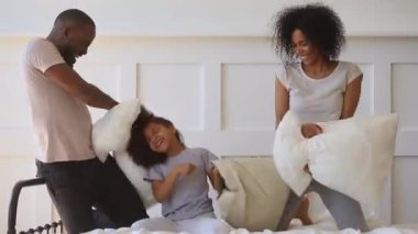African kid daughter playing pillow fight on bed with parents