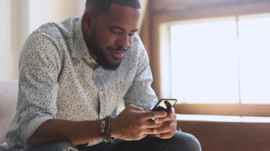 Young african man holding smartphone texting message sitting at home