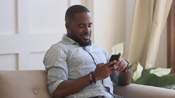 Smiling african american man holding smartphone texting at home — Stockvideo