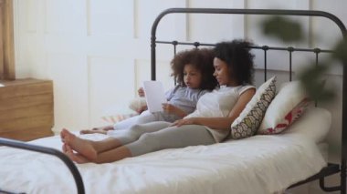 Focused african child daughter reading book on bed with mother