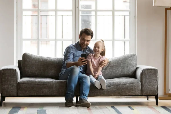 Happy father with little daughter using smartphone, sitting on couch — Stok fotoğraf