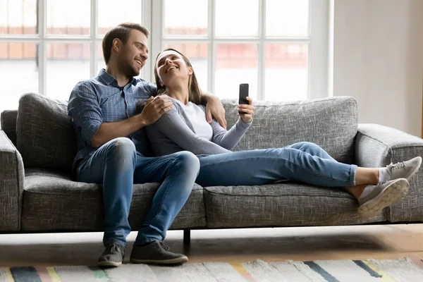 Happy young couple hugging, relaxing on couch, using smartphone — Stok fotoğraf