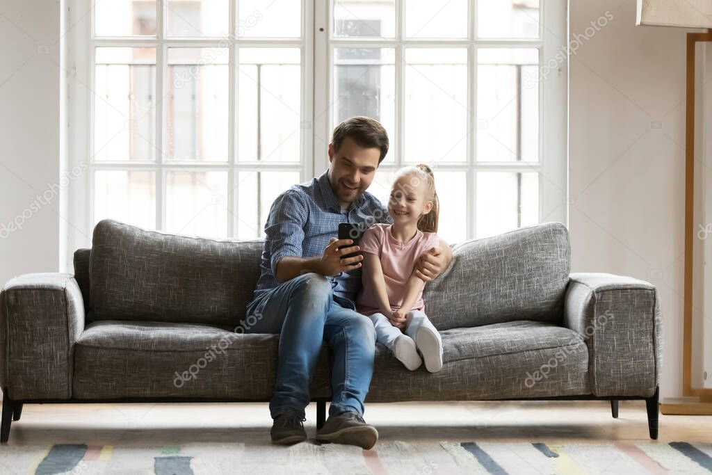Happy father with little daughter using smartphone, sitting on couch