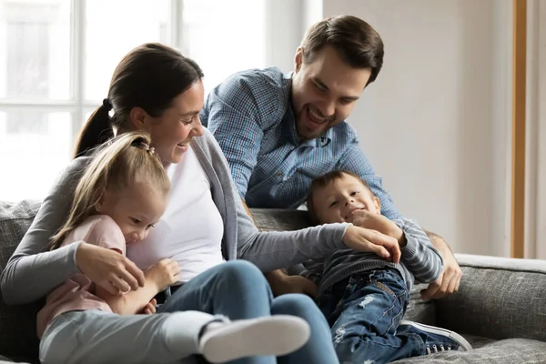 Счастливая молодая семья с маленькими детьми щекочущими на диване — стоковое фото