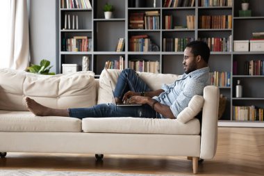 African guy lying on sofa typing message using computer
