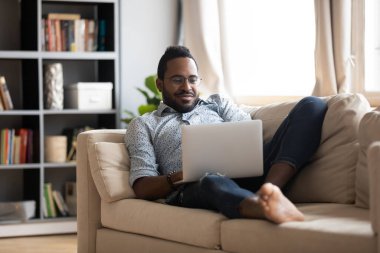 African guy lying on couch use notebook chatting with friends