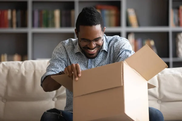 African guy opening parcel box seated on couch at home — Stockfoto