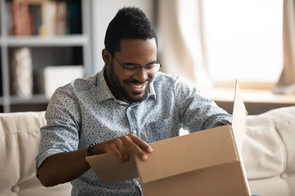 African man opening parcel seated on couch at home — Stockfoto