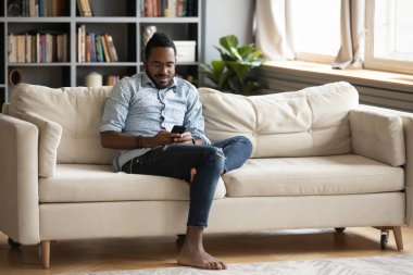 African man resting on couch holding smartphone chatting with friend