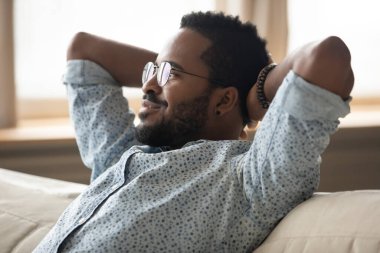 Calm African guy puts hands behind head relaxing on couch