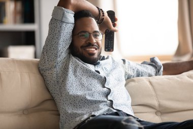 African guy resting on couch holding remote control watching tv