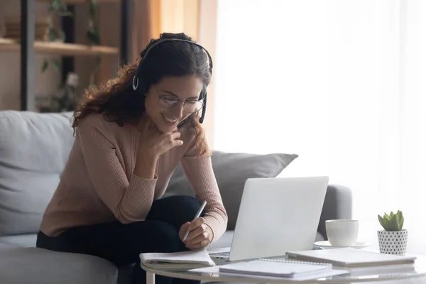 Mulher sorridente usando fones de ouvido escrevendo notas, estudando online — Fotografia de Stock