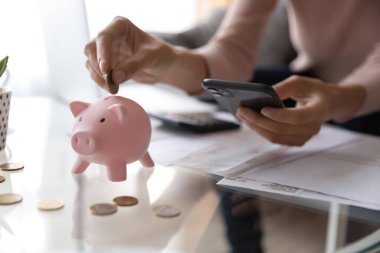 Close up woman putting coin in piggy bank, using smartphone