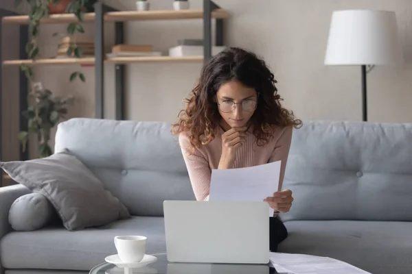 Thoughtful woman wearing glasses reading document, using laptop — Stockfoto