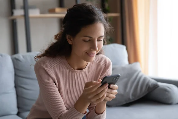 Smiling young woman using smartphone, sitting on cozy couch — Stock fotografie