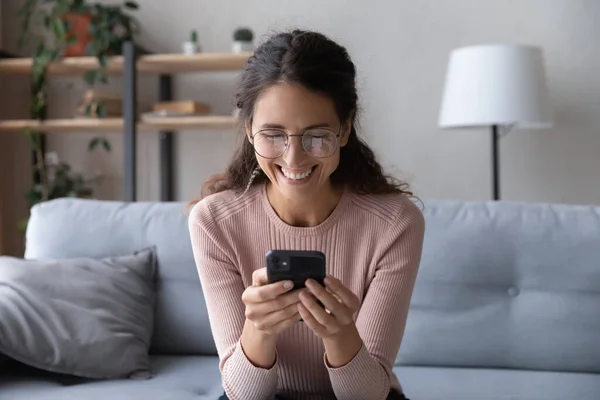 Smiling woman wearing glasses looking at smartphone screen, having fun — Stok fotoğraf