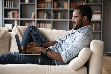African fellow lying on sofa typing message using laptop
