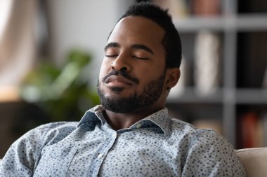 Close up African young man sit on couch closed eyes resting reduces fatigue