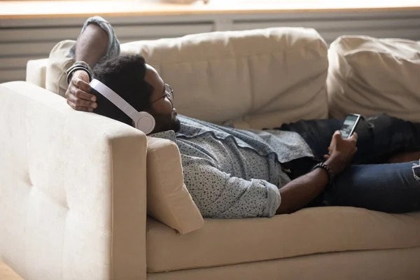 Guy resting on couch wearing headphones holding smartphone listening music