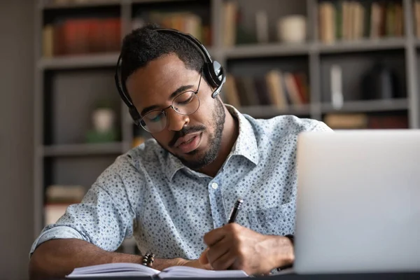 Focused african student writing notes listening lecture studying online