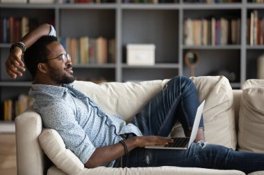 African guy relieving fatigue resting on comfy couch with notebook