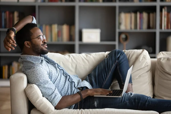 African guy relieving fatigue resting on comfy couch with notebook — Stok fotoğraf
