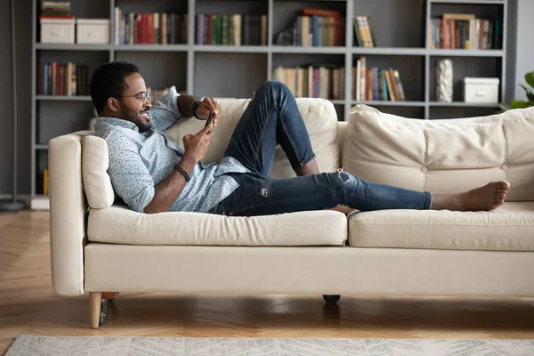 African guy lie on sofa with smartphone websurfing at home — Stock fotografie