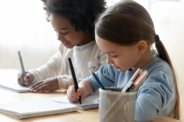 African and Caucasian schoolgirl mates sit at table writing task