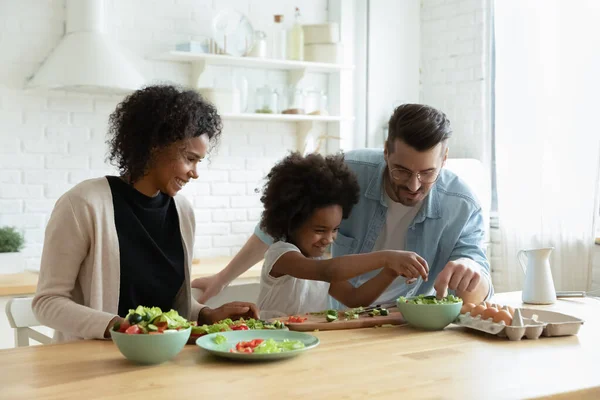 Caring multiethnic couple teach small daughter cook vegetable salad — Stockfoto