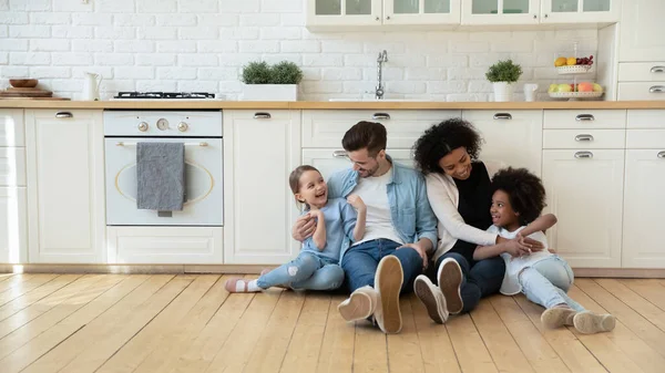 Proprietários multinacionais família com filhas sentadas no chão da cozinha moderna — Fotografia de Stock