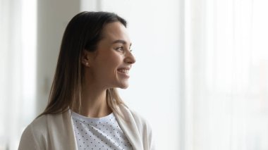 Pencerenin yanında duran mutlu genç kadın uzaktan bakıp gülümsüyor.