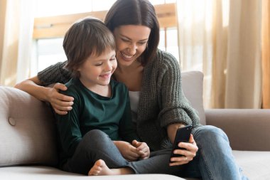 Mutlu anne ve küçük oğlu akıllı telefonu birlikte kullanıyor, kanepede oturuyor.