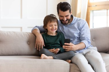 Mutlu baba ve küçük oğlu akıllı telefon kullanıyor, kanepede oturuyor.