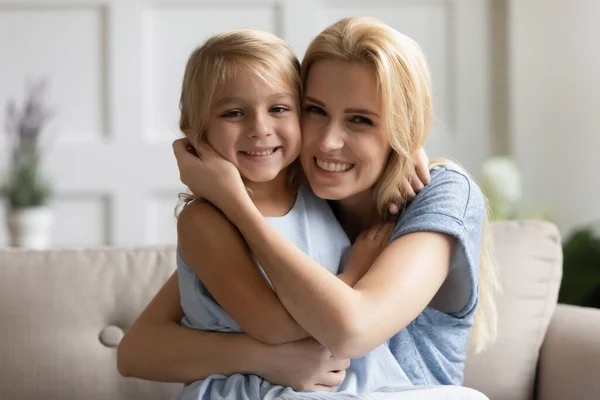 Mother cuddles daughter sitting on sofa smiling looking at camera