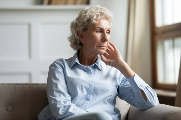 Concerned elderly woman lost in sad thoughts sitting on couch