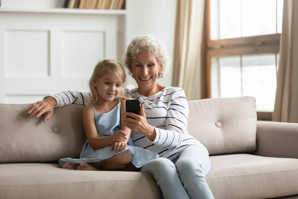 Старая бабушка маленькая внучка сидит на диване со смартфоном — стоковое фото
