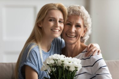 Yaşlı anne, yetişkin kız elinde çiçeklerle kanepede oturuyor.