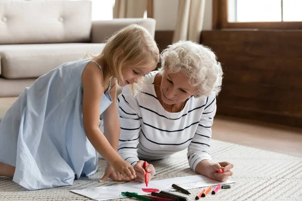 Ältere Oma bringt kleiner Enkelin Zeichnen auf warmem Boden bei — Stockfoto